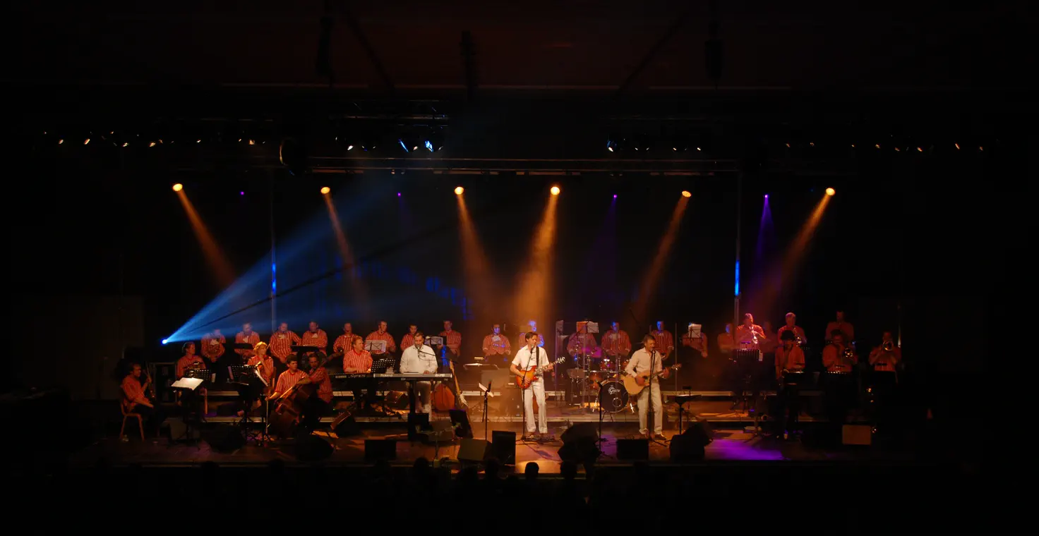 Leuster op het podium met een orkest en een koor in Urker streepbloezen, in blauw en oranje podiumlicht, voor een volle zaal.
